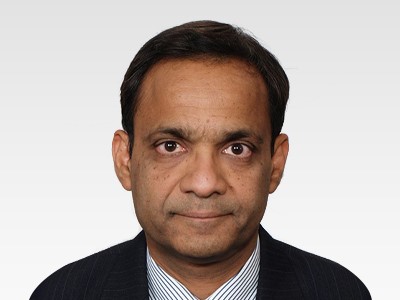 Photograph of Dr Alok Aggarwal wearing a black blazer over a white shirt with green stripes. He has a pleasent smile, bright eyes and hair combed sideways from left to right.