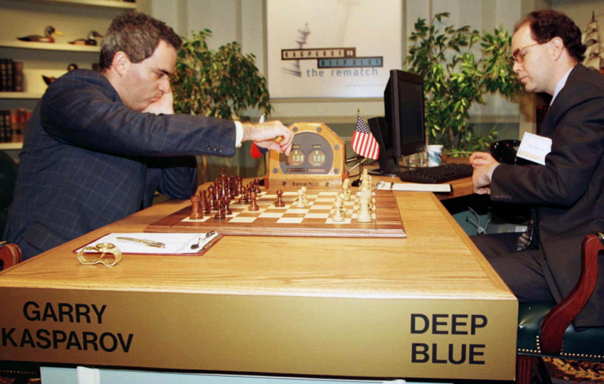 Picture of chess player Garry Kasparov seated facing a chess board with 16 chess pieces on one side and 16 chess pieces on the opposite side. He is competing with the IBM supercomputer named deep blue. The computer is monitored by a man seated facing a desktop computer wearing a black coat and taking notes in a notepad. Garry Kasparov wearing a checked black blazer over white shirt is moving a chess piece with his right arm and left arm placed on his cheek and elbow rested on the table